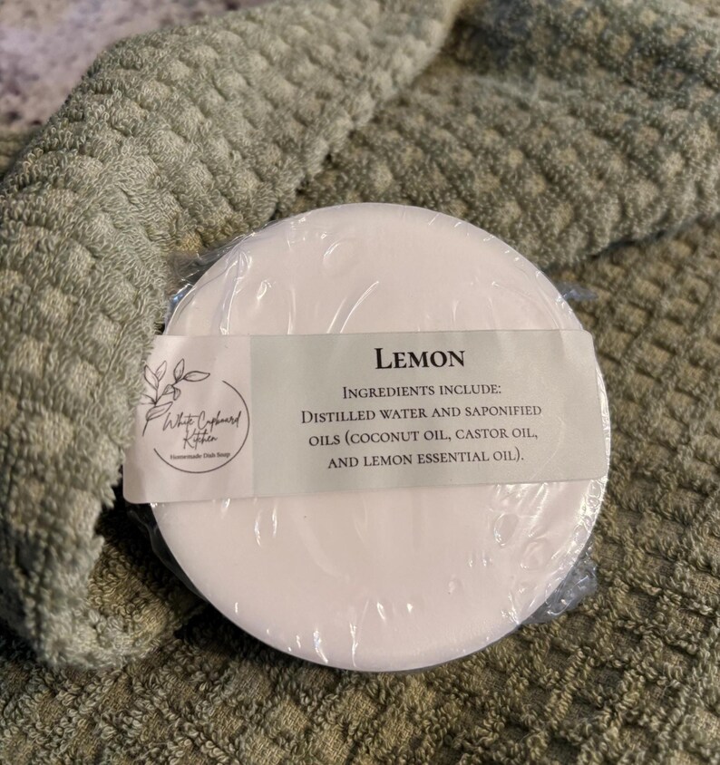 May include: A round, white bar of soap wrapped in clear plastic with a label that reads Lemon, Ingredients include: Distilled water and saponified oils (coconut oil, castor oil, and lemon essential oil), and a White Cupboard Kitchen logo.