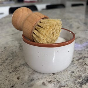 May include: A round wooden dish brush with light tan bristles resting inside a small white ceramic bowl. The bowl features a speckled texture and a thin brown rim around the top edge, containing a white substance at the bottom of the container.