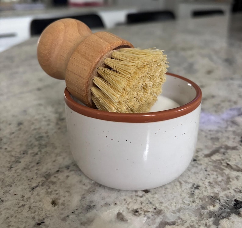 May include: A round wooden dish brush with light tan bristles resting inside a small white ceramic bowl. The bowl features a speckled texture and a thin brown rim around the top edge, containing a white substance at the bottom of the container.