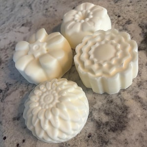 May include: Four round, off-white soap bars with distinct molded floral patterns on their tops. Each bar features a unique embossed design, including petal-like textures and geometric shapes, arranged in a cluster on a speckled surface.