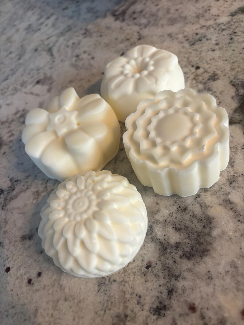 May include: Four round, off-white soap bars with distinct molded floral patterns on their tops. Each bar features a unique embossed design, including petal-like textures and geometric shapes, arranged in a cluster on a speckled surface.