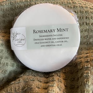 May include: A round white bar of Rosemary Mint soap wrapped in clear plastic. A label features the text White Cupboard Kitchen, Rosemary Mint, and a list of ingredients including distilled water, saponified coconut oil, castor oil, and essential oils.