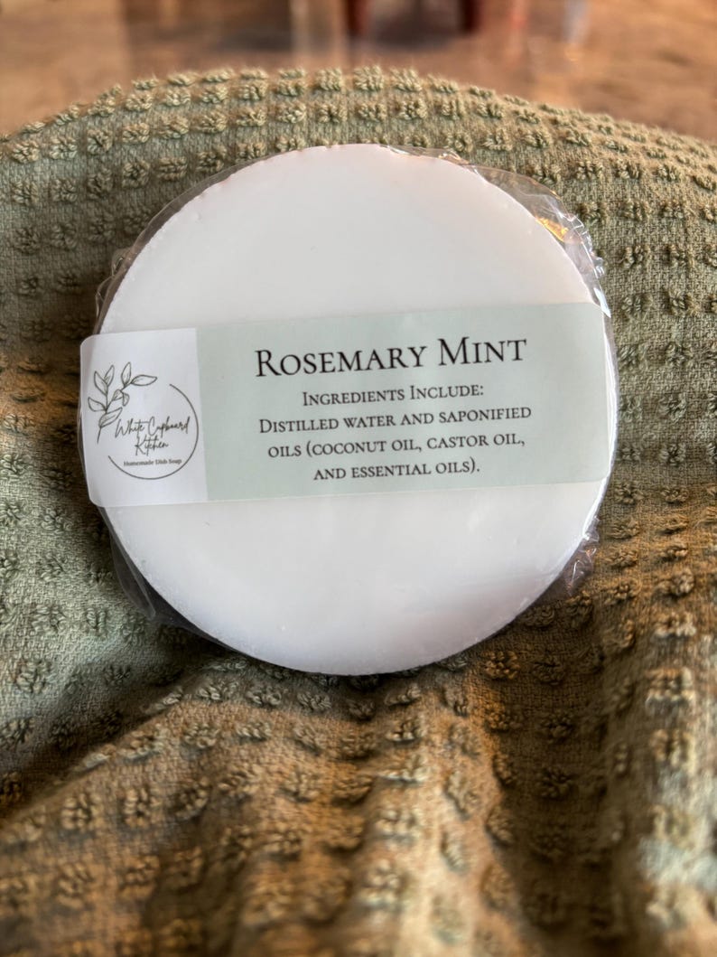 May include: A round white bar of Rosemary Mint soap wrapped in clear plastic. A label features the text White Cupboard Kitchen, Rosemary Mint, and a list of ingredients including distilled water, saponified coconut oil, castor oil, and essential oils.