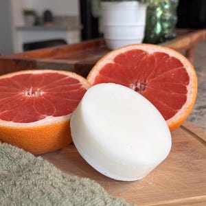 May include: A round, solid white bar of soap positioned in front of two halved grapefruits. The grapefruits display a vibrant pinkish-red interior flesh with a textured orange rind. The items are arranged together on a wooden surface.