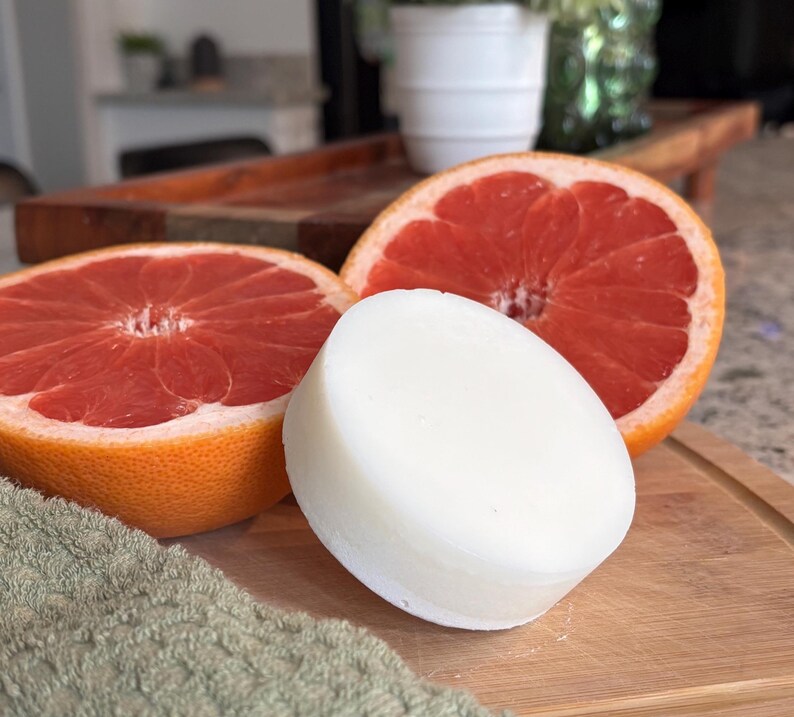 May include: A round, solid white bar of soap positioned in front of two halved grapefruits. The grapefruits display a vibrant pinkish-red interior flesh with a textured orange rind. The items are arranged together on a wooden surface.