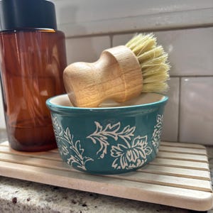 May include: A teal ceramic bowl with a white floral pattern, a wooden dish brush with light-colored bristles, and an amber glass soap dispenser with a black pump, all resting on a light-colored wooden slatted tray for kitchen organization.