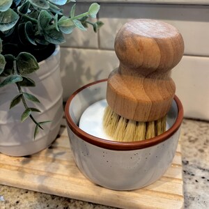 May include: A round wooden dish brush with light-colored natural bristles sits inside a small, speckled white ceramic bowl with a brown rim. The brush features a smooth, ergonomic wooden handle designed for a comfortable grip during cleaning tasks.