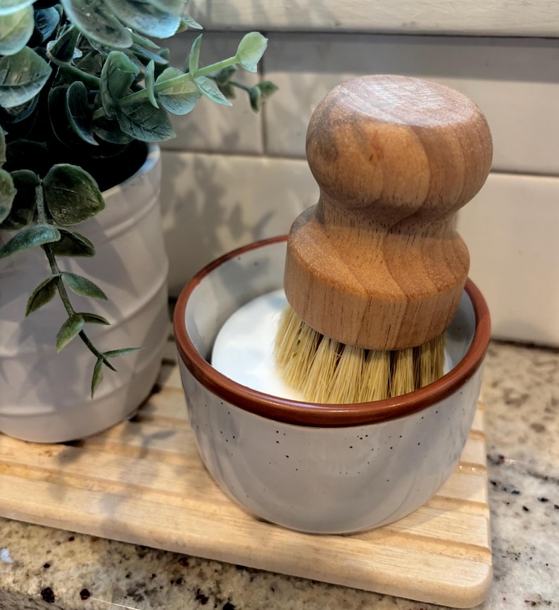 May include: A round wooden dish brush with light-colored natural bristles sits inside a small, speckled white ceramic bowl with a brown rim. The brush features a smooth, ergonomic wooden handle designed for a comfortable grip during cleaning tasks.