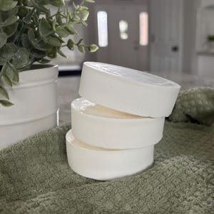 May include: Three round, solid white bars of soap stacked vertically on top of each other. The soap bars have a smooth, matte finish and a cylindrical shape. The stack is positioned on a textured, sage green fabric surface with a white planter nearby.