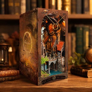 Può includere: Un diorama dettagliato e illuminato di una scena di villaggio fantasy, racchiuso in una cornice a forma di libro. La scena presenta edifici in miniatura, ponti e un fiume, con un anello d'oro sospeso dall'alto. Il libro recita "The Lord of the Rings".
