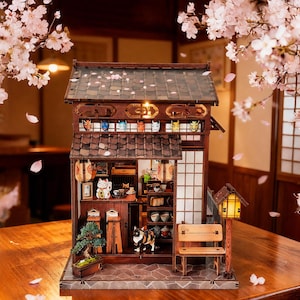 Könnte beinhalten: Ein detailliertes Holzmodell eines japanischen Hauses mit Ziegeldach, Papierlaternen und einer kleinen Bank. Der Innenraum zeigt Miniaturmöbel, Figuren und einen Bonsai-Baum. Kirschblüten umrahmen die Szene.