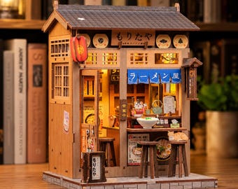 May include: A miniature wooden Japanese ramen shop model with intricate details. The model features a tiled roof, paper lanterns, and signage in Japanese. The interior is lit, showcasing a small dining area with miniature tables, chairs, and food items.