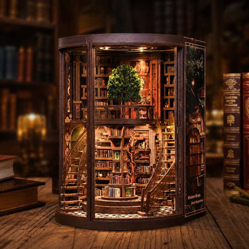Op de afbeelding: Gedetailleerd houten boekenplank-diorama met een miniatuurboom, wenteltrappen en binnenverlichting. Het diorama bevat talloze kleine boeken en decoratieve elementen, waardoor een grillige, gedetailleerde sc&egrave;ne ontstaat. De tekst "Wisdom Rings Book Repository" is zichtbaar.