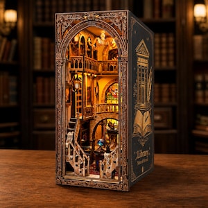 May include: A book nook diorama featuring a miniature library interior with warm lighting, staircases, balconies, and a stained glass window. The exterior side panel is black with gold illustrations of a building, a quill, and the text Library Notes of the soul.