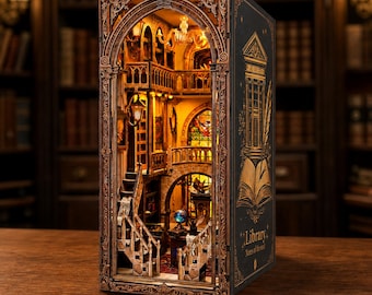 Puede incluir: Un diorama book nook que muestra el interior de una biblioteca en miniatura con iluminación cálida, escaleras, balcones y una vidriera. El panel lateral exterior es negro con ilustraciones doradas de un edificio, una pluma y el texto Library Notes of the soul.