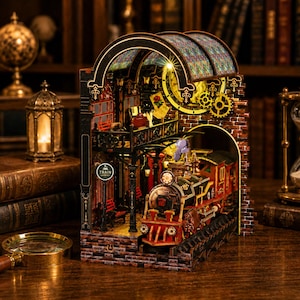 May include: A detailed, multi-layered wooden model of a train station with a steam train. The model features a brick facade, arched windows, and a clock tower. The word "TRAIN" is visible on a sign. The train is red and black.