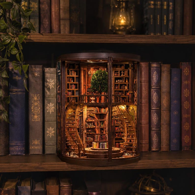 Op de afbeelding: Een gedetailleerd houten boekenplank-diorama met een miniatuur interieur bibliotheeksc&egrave;ne. Het diorama heeft wenteltrappen, boekenplanken en een kleine boom. De sc&egrave;ne is verlicht, waardoor een warme gloed ontstaat. Het diorama staat op een houten plank met boeken.