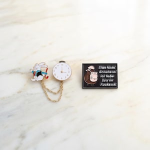 Puede incluir: Tres broches de esmalte: un conejo blanco corriendo con un atuendo rojo y azul, un reloj blanco con ribete dorado y un pin negro con un pájaro y texto en alemán. El reloj y el conejo están conectados por una cadena dorada.
