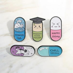 Può includere: Una collezione di cinque spille smaltate colorate a forma di pillola. I disegni includono un'onda con "Vitamin Sea", un laureato con "Pharmacy School Survivor", una pillola sorridente con "Happy 250mg", un gatto e una pillola blu con "Chill 1000mg".