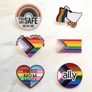 May include: A collection of colorful enamel pins with LGBTQ+ pride themes. Designs include a rainbow, a goose holding a rainbow flag, a heart with the text "Free Mom Hugs", and pins with the text "You are safe with me" and "ally".