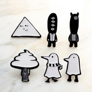 Puede incluir: Colección de broches de esmalte con diseños caprichosos y de dibujos animados. Los broches incluyen un triángulo blanco con una cara, varias figuras en blanco y negro y un personaje parecido a un pingüino con una bufanda. Los broches tienen contornos negros.
