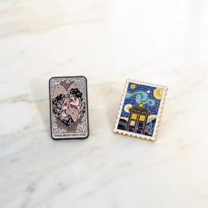 May include: Two enamel pins. One is a rectangular pin with a couple kissing, with the text "THINGS ARE BETTER IF I STAY". The other is a postage stamp pin with a "Police Box" design and a starry night background.