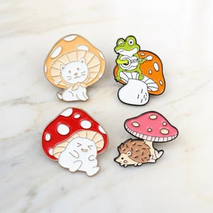 Può includere: Quattro spille smaltate con disegni di funghi. Un gatto bianco sotto un fungo arancione. Una rana che suona la chitarra su un fungo. Una creatura bianca sorridente sotto un fungo rosso. Un riccio sotto un fungo rosa.