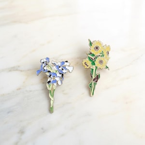 Puede incluir: Dos broches de flores de esmalte. Uno presenta un ramo de iris azules y blancos con tallos verdes. El otro muestra un grupo de girasoles amarillos con hojas y tallos verdes. Ambos broches tienen un contorno dorado.