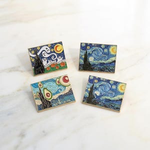 May include: Four square enamel pins featuring art-inspired designs. One pin depicts a Halloween scene with ghosts and pumpkins. The other three pins feature a starry night and avocado designs. Each pin has a gold border.