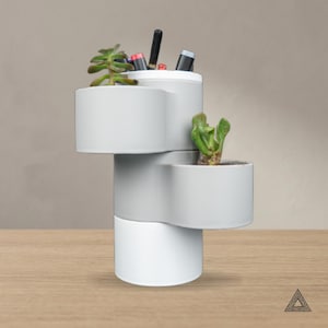 Pode incluir: Um organizador de secretária cilíndrico de níveis em cinzento claro e branco, com três compartimentos circulares desalinhados. A secção superior contém canetas e uma pequena suculenta verde, enquanto a secção central contém outra suculenta.