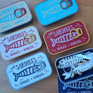 Könnte beinhalten: Eine Sammlung rechteckiger Metalldosen mit Aufreißdeckeln, die jeweils ein Fischmotiv sowie den Text "not SARDINES" und "SKINLESS + BONELESS" zeigen. Eine geöffnete Dose enthält einen weißen fischförmigen Einsatz mit verschiedenen Schmuckstücken.