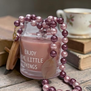 以下が含まれることがあります： ピンクと紫のビーズのネックレス、大きなペンダント付き。 ネックレスは「ENJOY THE LITTLE THINGS」の文字が入ったキャンドルジャーの上にかけられています。 背景にはティーカップと本があり、木製の表面に置かれています。