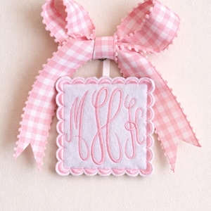 May include: A pink and white gingham ribbon bow tops a square, scalloped-edged ornament. The ornament features a white center with pink embroidered initials. The overall design is soft and feminine.