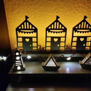 May include: A collection of decorative wooden light-up houses and boats. The houses have a black silhouette design with heart cutouts and a flag on top. The boats and lighthouse are also illuminated, with a string of star-shaped lights.