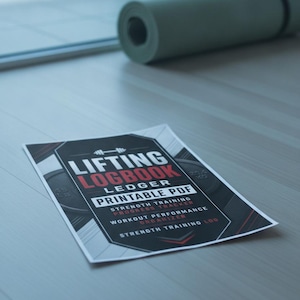 May include: A rectangular paper sheet featuring the text LIFTING LOGBOOK LEDGER PRINTABLE PDF, STRENGTH TRAINING PROGRESS TRACKER, WORKOUT PERFORMANCE ORGANIZER, and STRENGTH TRAINING LOG in white and red against a black and gray geometric background.
