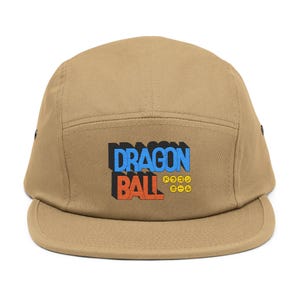 May include: Tan five-panel cap featuring the text "DRAGON BALL" in blue and orange, with Japanese text and symbols below. The cap has a flat brim and a structured crown.