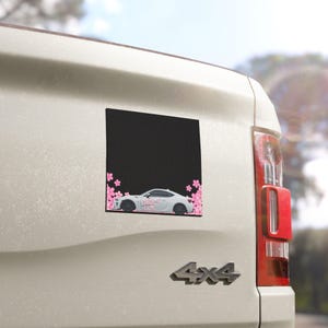 May include: A black square magnet with a white sports car design and pink cherry blossom accents. The car is depicted in profile, with the blossoms framing the bottom and sides of the black background.