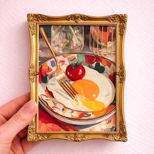 Puede incluir: Pintura enmarcada de una escena de desayuno. La obra presenta un huevo frito con una yema de color naranja brillante, un tomate rojo y un tenedor sobre un plato con un diseño floral colorido. El marco es dorado con detalles ornamentados.