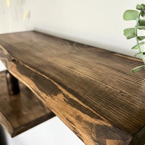 Könnte beinhalten: Nahaufnahme eines rustikalen, dunkelbraunen Holzregals mit einer Baumkante. Das Regal weist sichtbare Holzmaserungen und Knoten auf, was ihm ein natürliches, strukturiertes Aussehen verleiht. Das Regal ist an einer Wand befestigt, und ein kleineres Regal ist darunter sichtbar.