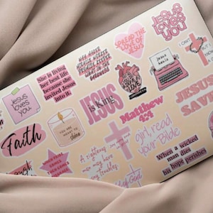 May include: A collection of pink and white stickers with religious themes and phrases, including "Jesus loves you," "Faith," and Bible verses. The stickers feature crosses, hearts, and other related imagery.