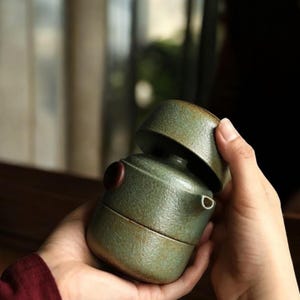 May include: A cylindrical ceramic teapot with a textured, mottled sage green finish. The teapot features a small, dark brown circular knob on the side, a short spout, and a matching lid that sits securely on top of the main body of the vessel.