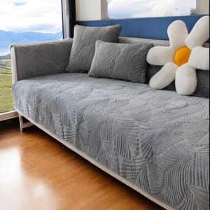 May include: A gray sofa cover featuring a textured leaf pattern, accompanied by two matching gray throw pillows and a large white flower-shaped cushion with a yellow center. The cover drapes over the seat and backrest of the sofa.