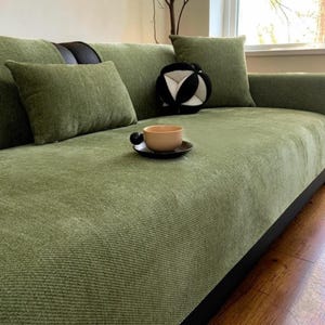 May include: A sage green textured sofa cover featuring a ribbed fabric finish, accompanied by matching rectangular and square throw pillows. A small, solid light brown ceramic cup rests on a dark saucer placed directly on the sofa seat.