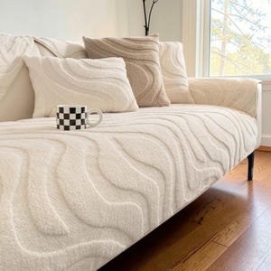 Può includere: Un copridivano color crema con un motivo ondulato testurizzato, completo di cuscini abbinati color crema e beige. Una tazza in ceramica con motivo a scacchi bianco e nero è appoggiata sulla seduta. Il tessuto ha un aspetto morbido e soffice.