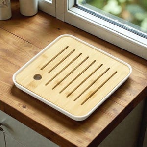 Può includere: Un vassoio da tè quadrato in bambù con bordo bianco. Il vassoio presenta una serie di scanalature parallele e un piccolo foro circolare. Il bambù è marrone chiaro e il bordo è bianco. Progettato per la preparazione del tè.