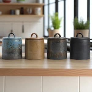 May include: Four cylindrical ceramic canisters with lids, each featuring a small arched handle on top. The canisters are arranged in a row and display distinct finishes: a blue and grey mottled glaze, a solid tan, a dark textured brown, and a matte black.