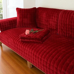 May include: A deep red sofa cover with a textured, grid-like square pattern. The cover features a matching square throw pillow and a folded piece of the same fabric resting on the seat, topped with a single artificial pink rose.