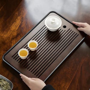May include: A dark brown wooden tea tray with a slotted design, holding two small white teacups filled with tea and a white lidded teapot. The rectangular tray has a black border. The tea set is designed for serving tea.