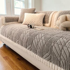 May include: A light grey sofa cover with a textured geometric pattern and white pom-pom trim edges. The cover is draped over a sofa, accompanied by a matching rectangular throw pillow, a square throw pillow, and a knot-shaped decorative pillow.