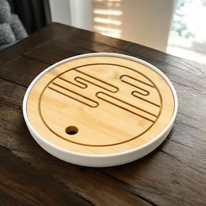 May include: A round tea tray with a white rim and a light brown bamboo center. The center features a carved design with a small hole. The tray sits on a dark wooden surface.